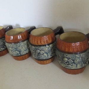 May include: Four brown ceramic mugs shaped like barrels with blue and white painted designs. The mugs have handles and are arranged in a row.