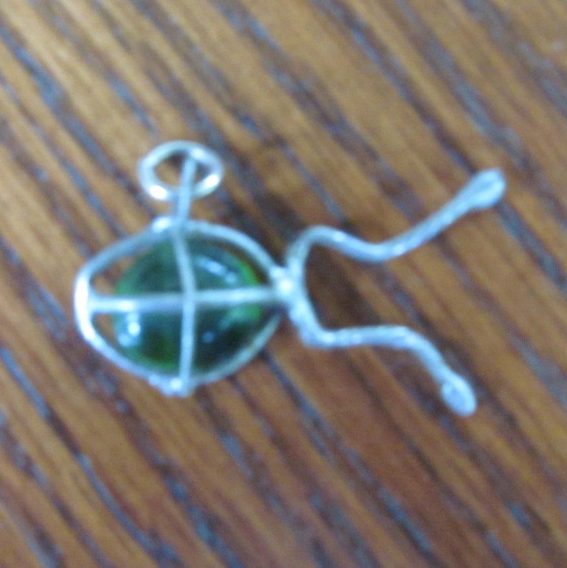 May include: A handmade pendant featuring a green glass orb encased in a silver-colored wire cage. The wire extends into two curved, decorative elements. The pendant is designed to be worn as jewelry.