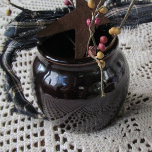 May include: A dark brown ceramic pot with a rustic star and berry sprigs inside. The pot is sitting on a white crocheted doily.