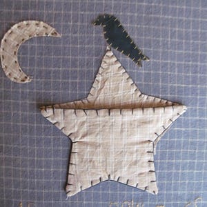 May include: A framed textile art piece featuring a beige star, crescent moon, and a dark blue bird. The star and moon are made of patterned fabric and stitched with dark thread. The background is a blue grid pattern.