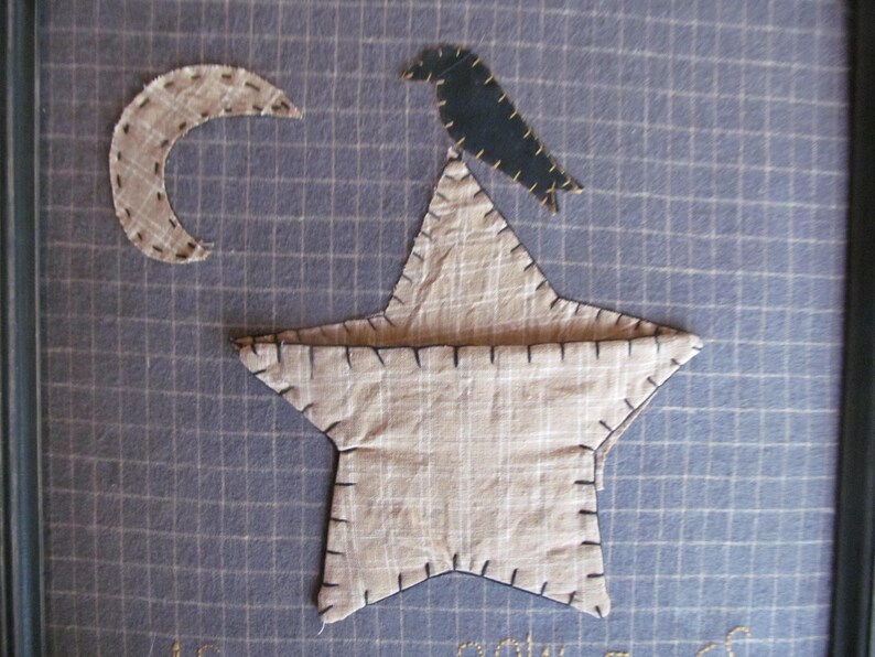 May include: A framed textile art piece featuring a beige star, crescent moon, and a dark blue bird. The star and moon are made of patterned fabric and stitched with dark thread. The background is a blue grid pattern.