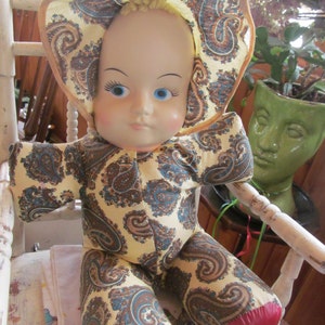 K&ouml;nnte beinhalten: Eine Vintage-Puppe mit einem Kleid im Paisley-Muster und einem rot-wei&szlig;en Hut. Die Puppe hat blaue Augen und ein bemaltes Gesicht.