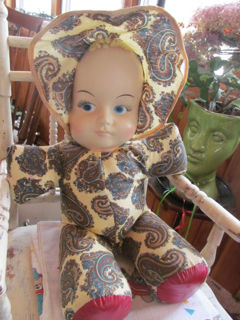K&ouml;nnte beinhalten: Eine Vintage-Puppe mit einem Kleid im Paisley-Muster und einem rot-wei&szlig;en Hut. Die Puppe hat blaue Augen und ein bemaltes Gesicht.