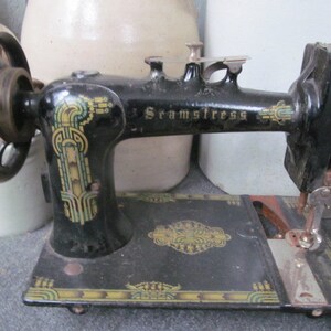Op de afbeelding: Een zwarte vintage naaimachine met een decoratief ontwerp aan de zijkant. De machine heeft een metalen voetpedaal en een naald die met wit garen is ingedraaid. De machine is gelabeld met "Seamstress".