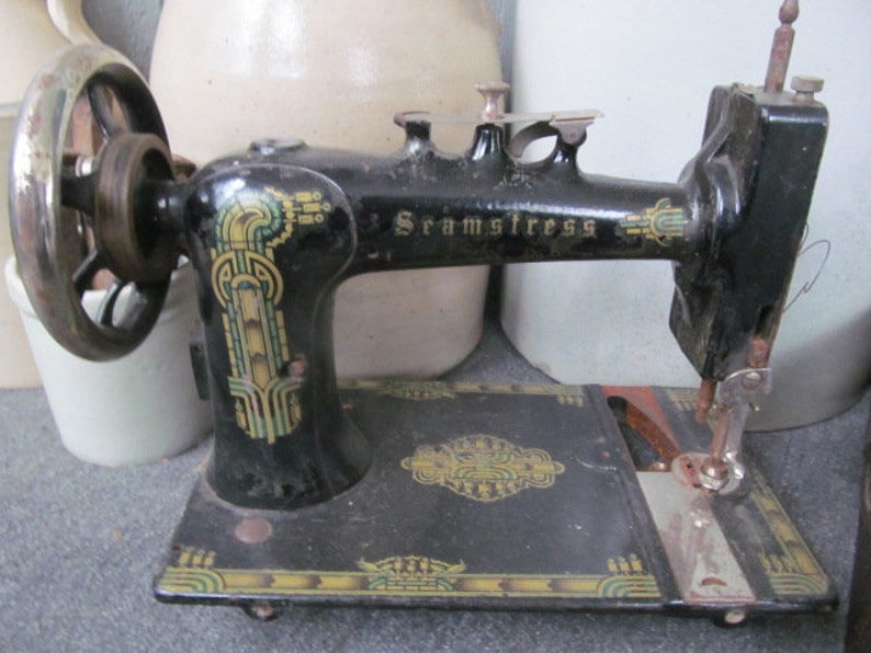 Op de afbeelding: Een zwarte vintage naaimachine met een decoratief ontwerp aan de zijkant. De machine heeft een metalen voetpedaal en een naald die met wit garen is ingedraaid. De machine is gelabeld met "Seamstress".