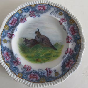 May include: A white porcelain plate with a blue and gold floral border. The centre of the plate features a hand-painted image of two quail in a grassy field.