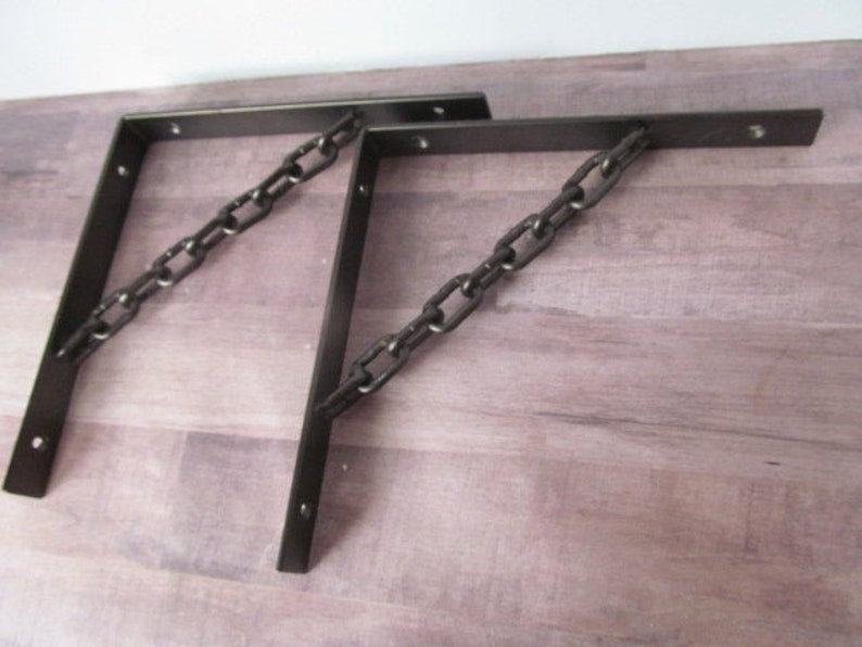 May include: Two black metal shelf brackets with a chain design. The brackets are attached to a wooden surface.