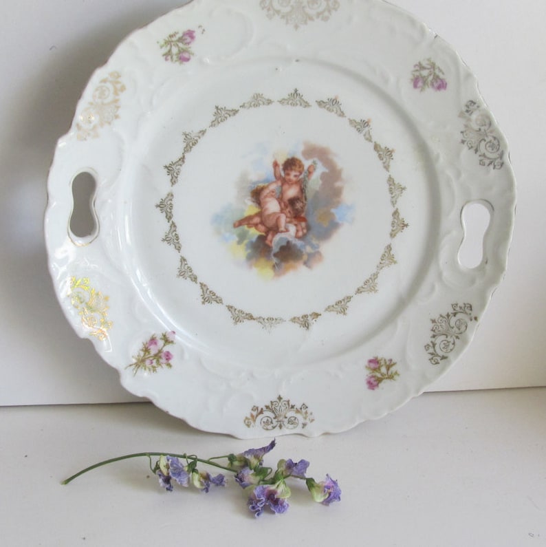 May include: White porcelain serving plate with two handles. The plate features a gold trim and a central image of two cherubs in the clouds. The plate is decorated with pink flowers.