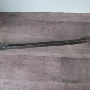 Puede incluir: Una palanca de metal negra con una garra en un extremo y un extremo bifurcado en el otro. La herramienta está sobre una superficie de madera marrón claro.