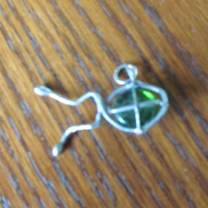 May include: A silver-toned pendant in the shape of a fish. The fish has a green, oval-shaped glass body with a silver wire frame. The pendant has a loop at the top for a chain. The fish's tail and fins are made of silver wire.