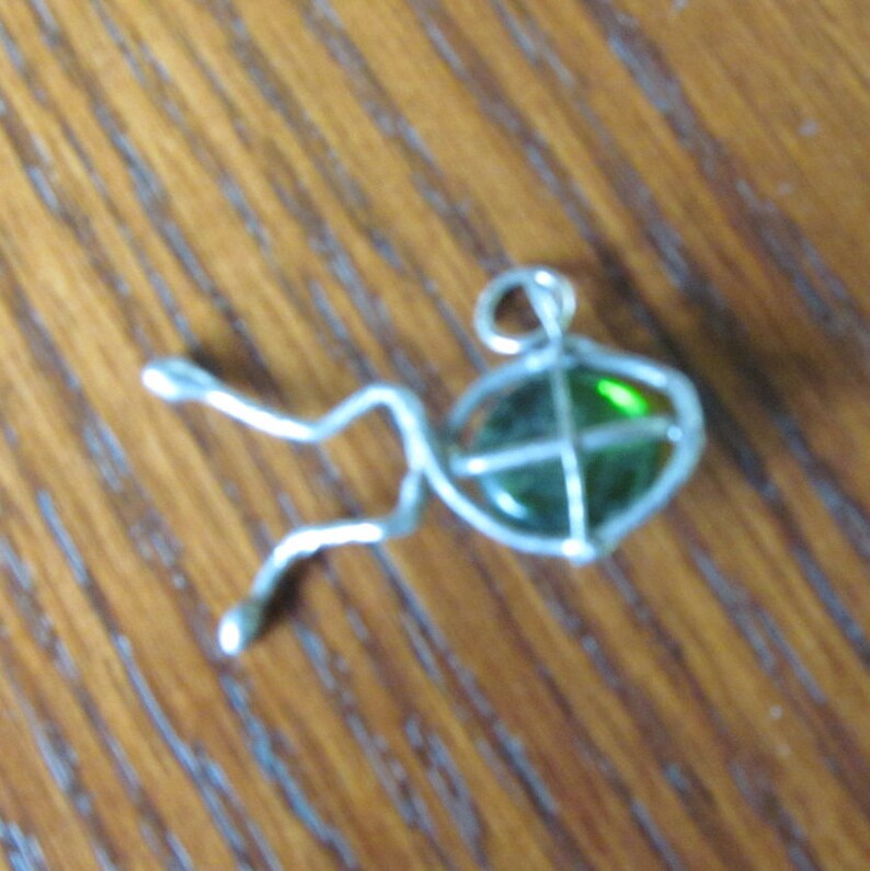 May include: A silver-toned pendant in the shape of a fish. The fish has a green, oval-shaped glass body with a silver wire frame. The pendant has a loop at the top for a chain. The fish's tail and fins are made of silver wire.