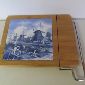 Op de afbeelding: Een houten kaasplank met een blauwe en witte tegel waarop windmolens en koeien zijn afgebeeld. De plank heeft een ingebouwde draad-kaassnijder.