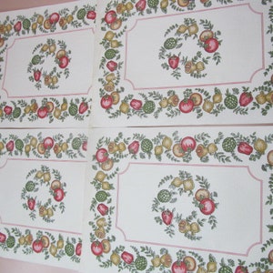 May include: Four white placemats with a red, green, and yellow floral border featuring a wreath of fruit and vegetables. The placemats are made of a fabric material.