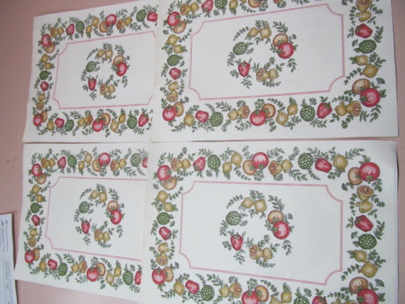 May include: Four white placemats with a red, green, and yellow floral border featuring a wreath of fruit and vegetables. The placemats are made of a fabric material.