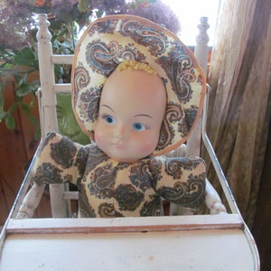 K&ouml;nnte beinhalten: Eine Vintage-Puppe mit bemaltem Gesicht und blauen Augen sitzt in einem wei&szlig;en Holz-Hochstuhl. Die Puppe tr&auml;gt ein gelbes und braunes Kleid und einen Hut mit Paisley-Muster.