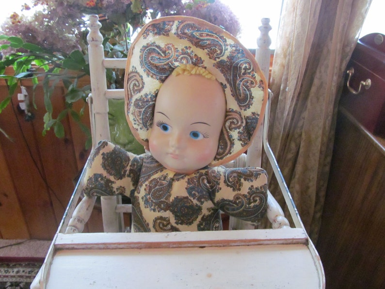 K&ouml;nnte beinhalten: Eine Vintage-Puppe mit bemaltem Gesicht und blauen Augen sitzt in einem wei&szlig;en Holz-Hochstuhl. Die Puppe tr&auml;gt ein gelbes und braunes Kleid und einen Hut mit Paisley-Muster.