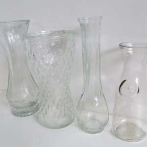 May include: Four clear glass vases of different shapes and sizes. The vases are all empty and are arranged on a white surface.