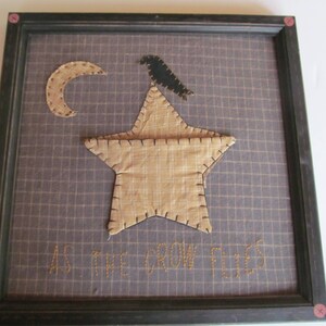 May include: Framed artwork featuring a tan star, crescent moon, and a black crow on a grid background. The phrase "AS THE CROW FLIES" is stitched below. The frame is dark with pink accents.