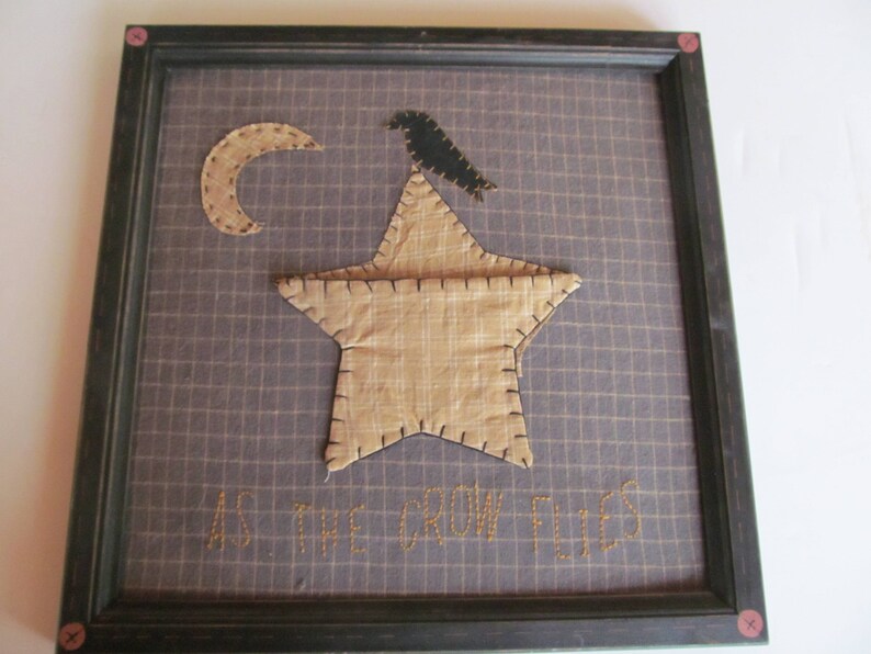 May include: Framed artwork featuring a tan star, crescent moon, and a black crow on a grid background. The phrase "AS THE CROW FLIES" is stitched below. The frame is dark with pink accents.