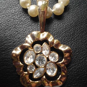 Pu&ograve; includere: Una collana con un ciondolo a forma di fiore dorato con dettagli in strass trasparenti. Il ciondolo &egrave; attaccato a una collana di perle.