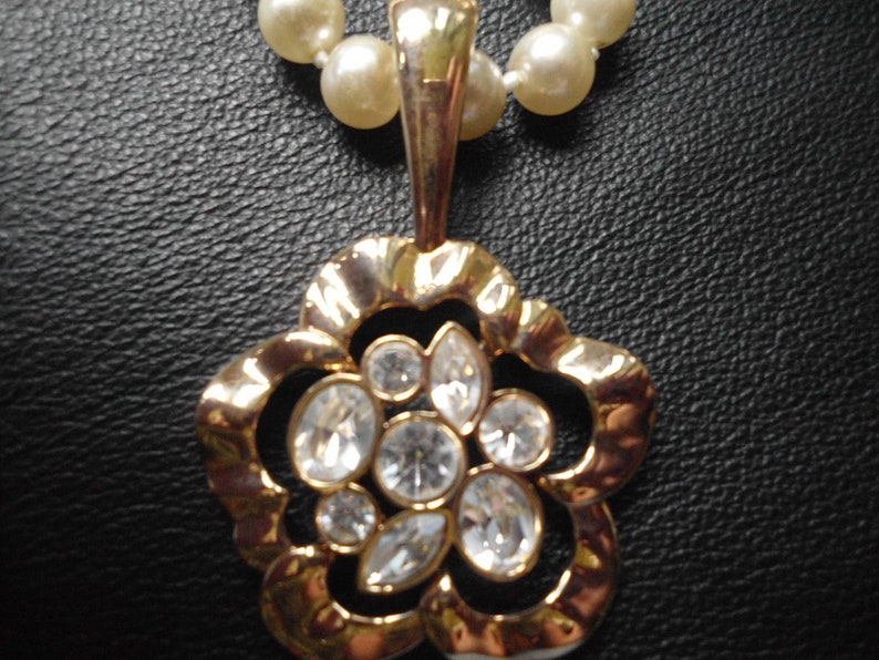 Pu&ograve; includere: Una collana con un ciondolo a forma di fiore dorato con dettagli in strass trasparenti. Il ciondolo &egrave; attaccato a una collana di perle.