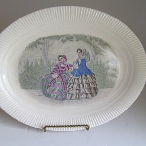 Puede incluir: Plato ovalado blanco para servir con un borde elevado y un diseño decorativo de dos mujeres con vestidos del siglo XIX de pie en un jardín.