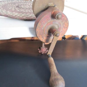 Puede incluir: Una rueda de afilar vintage de manivela con un mango de madera. La rueda es de metal y tiene un acabado rojo oxidado. El mango es de madera y tiene un acabado marrón oscuro.