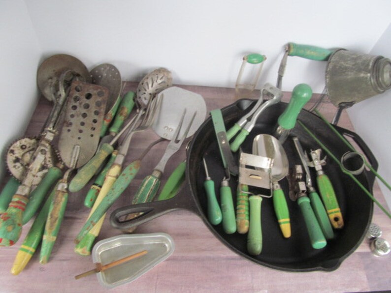 Puede incluir: Una colecci&oacute;n de utensilios de cocina vintage con mangos verdes. Los utensilios incluyen una variedad de herramientas, como un machacador de patatas, un batidor, una esp&aacute;tula, un rallador y una cuchara. Los utensilios est&aacute;n dispuestos en una sart&eacute;n de hierro fundido.
