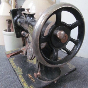 Op de afbeelding: Zwarte vintage naaimachine met een groot wiel en een decoratieve basis. De basis heeft gouden en groene accenten en de tekst "The Singer Manufacturing Co.  New York, U.S.A."