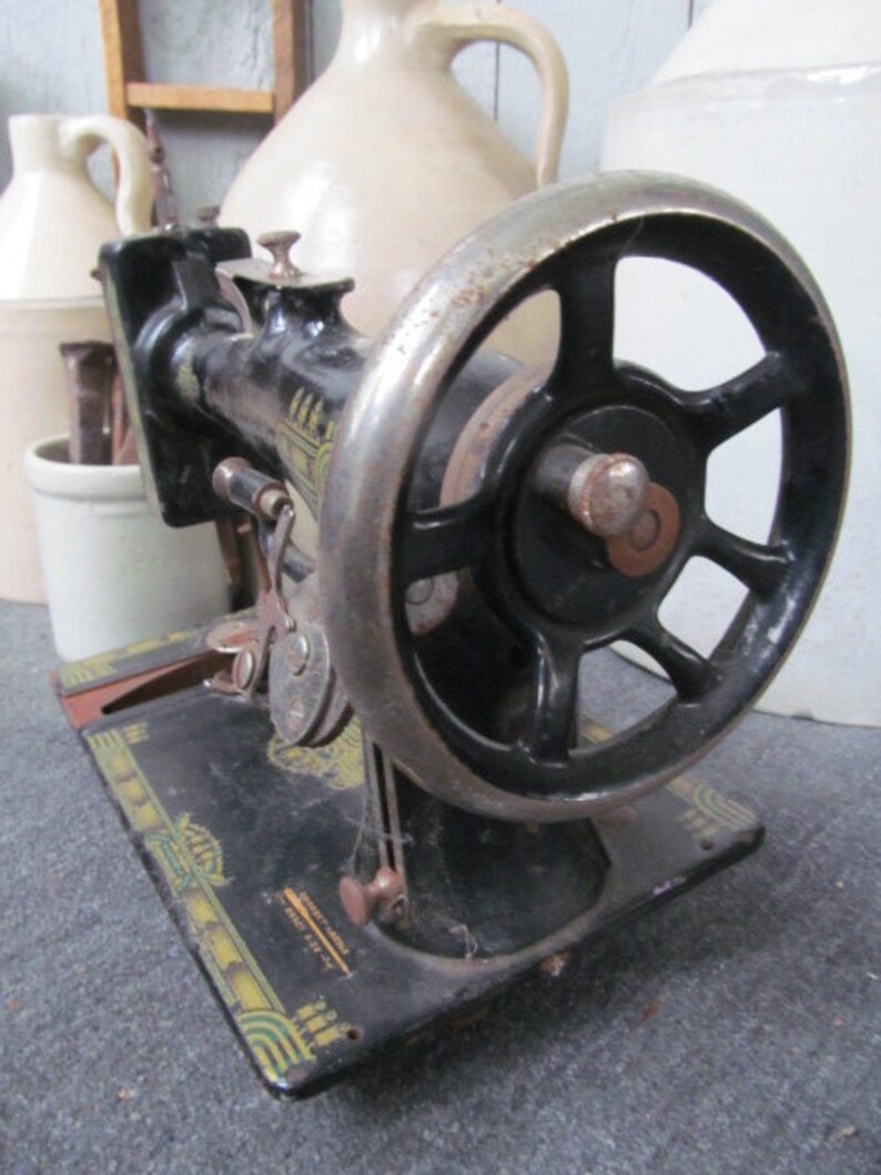 Op de afbeelding: Zwarte vintage naaimachine met een groot wiel en een decoratieve basis. De basis heeft gouden en groene accenten en de tekst "The Singer Manufacturing Co.  New York, U.S.A."