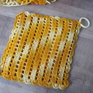 May include: A handmade, square crochet potholder in shades of yellow and cream. The potholder features a scalloped edge and a white ring for hanging. The open weave design allows for air circulation. The potholder is approximately 8 inches square.