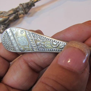 Op de afbeelding: Close-up van een antieke zilveren lepel met ingewikkelde gegraveerde patronen en een monogram-initiaal. De lepelsteel heeft een gedetailleerde figuur. De lepel wordt in een hand gehouden.