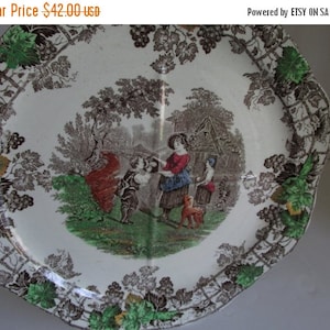 Könnte beinhalten: Ein antiker ovaler Servierteller aus Keramik mit einem braunen, grünen und weißen Blumendesign. Die Mitte des Tellers zeigt eine Szene mit Menschen in ländlicher Umgebung.