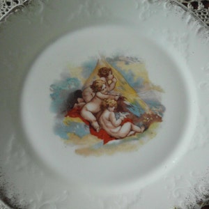 May include: A white porcelain plate with a gold rim and a decorative design of four cherubs in a cloud-like setting. The cherubs are painted in shades of pink, yellow, and blue.