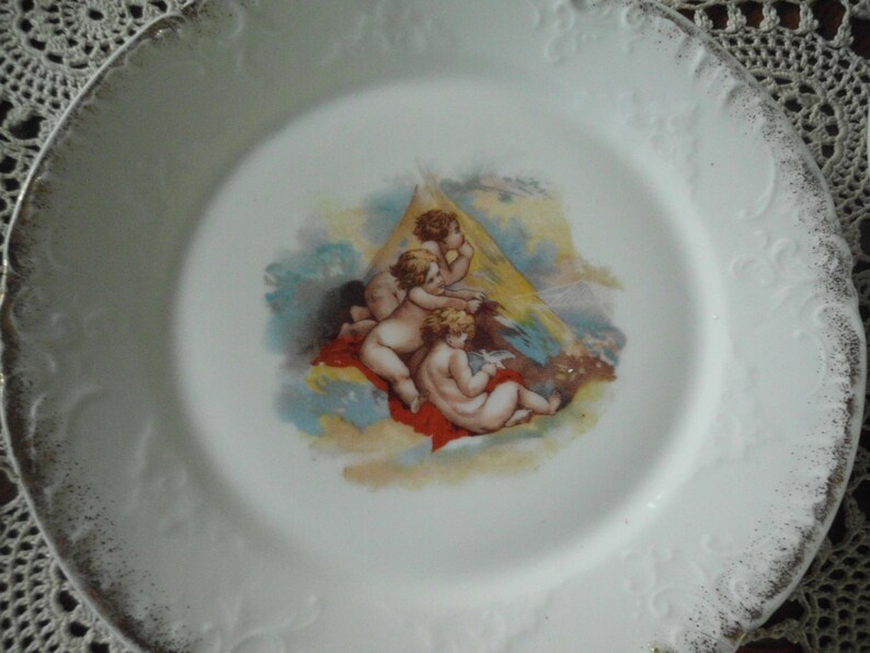 May include: A white porcelain plate with a gold rim and a decorative design of four cherubs in a cloud-like setting. The cherubs are painted in shades of pink, yellow, and blue.