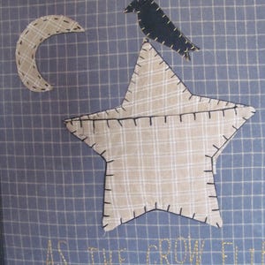 May include: Framed textile art featuring a star, crescent moon, and crow appliqued on a blue grid background. The star and moon are plaid, and the crow is black. The phrase "AS THE CROW FLIES" is embroidered below.