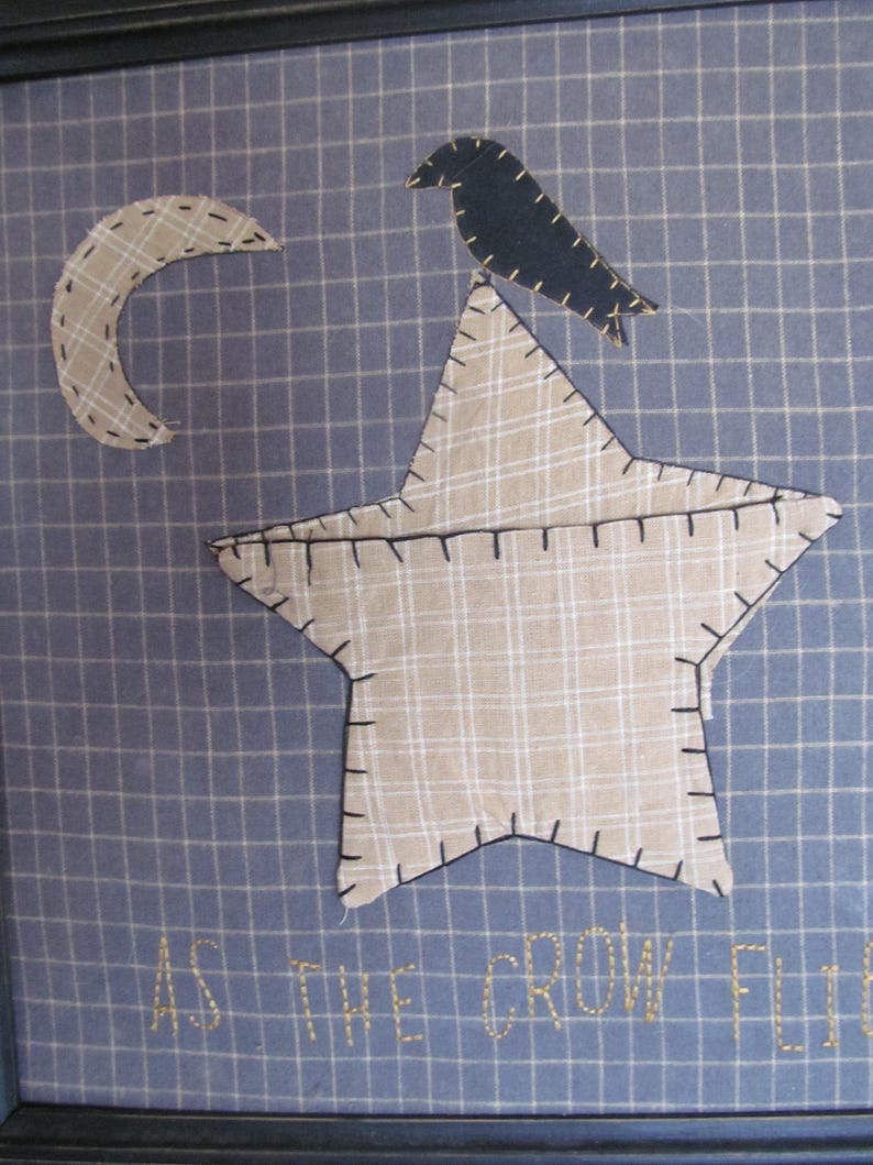 May include: Framed textile art featuring a star, crescent moon, and crow appliqued on a blue grid background. The star and moon are plaid, and the crow is black. The phrase "AS THE CROW FLIES" is embroidered below.