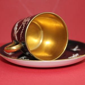 May include: A black teacup and saucer set with gold trim and a gold interior. The cup and saucer feature a white bird design. The cup is resting on the saucer, with the interior of the cup facing upwards.