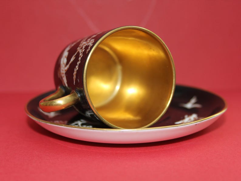May include: A black teacup and saucer set with gold trim and a gold interior. The cup and saucer feature a white bird design. The cup is resting on the saucer, with the interior of the cup facing upwards.