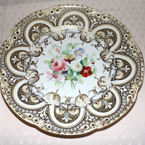 May include: An antique porcelain plate with a scalloped edge. The plate features a central floral bouquet in shades of pink, red, and white. The border has an ornate design in gold, grey, and white, creating an elegant aesthetic.