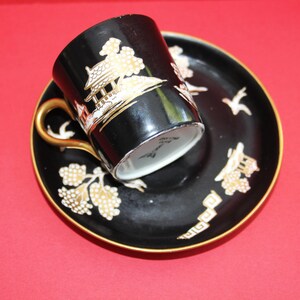 May include: Black teacup and saucer set with gold detailing. The cup and saucer feature a gold Asian-inspired landscape design. The cup has a gold handle and the saucer rim is gold. The set is on a red background.