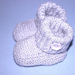 Puede incluir: Un par de botines para bebé tejidos en color gris claro con un botón de flor morado en un botín.