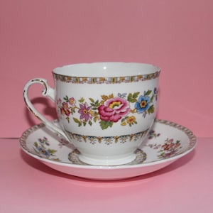May include: White teacup and saucer set with a floral design featuring pink, blue, and yellow flowers. The cup has a gold handle and trim. The saucer has a matching floral pattern.