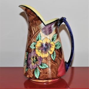May include: A brown ceramic jug with a yellow rim and blue handle. The jug is decorated with a floral pattern of purple and yellow pansies with green leaves.