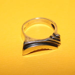 Peut inclure: Une bague en argent au design unique. La bague pr&eacute;sente un anneau lisse et incurv&eacute; qui s'&eacute;largit en une face plate et rectangulaire avec des rainures horizontales. La bague est pos&eacute;e sur un fond jaune.