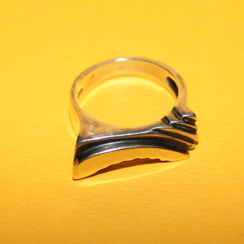 Peut inclure: Une bague en argent au design unique. La bague pr&eacute;sente un anneau lisse et incurv&eacute; qui s'&eacute;largit en une face plate et rectangulaire avec des rainures horizontales. La bague est pos&eacute;e sur un fond jaune.
