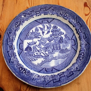 Può includere: Un piatto in porcellana blu e bianco con un design cinese tradizionale. Il piatto presenta un salice piangente, una pagoda e una barca. Il design è circondato da un bordo blu.