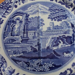 May include: Blue and white ceramic plate with a landscape scene. The plate features a detailed illustration of a rural landscape with buildings, trees, and figures. The rim has a floral and scroll design. A decorative piece for serving or display.