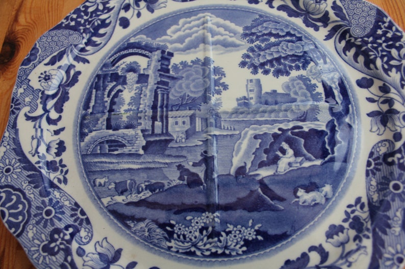 May include: Blue and white ceramic plate with a landscape scene. The plate features a detailed illustration of a rural landscape with buildings, trees, and figures. The rim has a floral and scroll design. A decorative piece for serving or display.