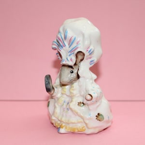 Puede incluir: Figura de cerámica de un ratón vestido con un atuendo rococó blanco y amarillo, sosteniendo un pequeño objeto oscuro. El ratón lleva un gran sombrero blanco con detalles azules y rosas. La figura está pintada a mano.